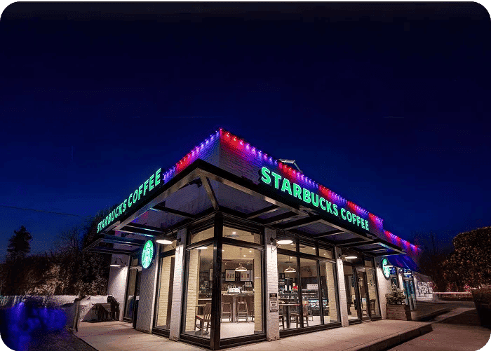 Starbucks coffee shop illuminated at night with colorful lights and glass exterior.