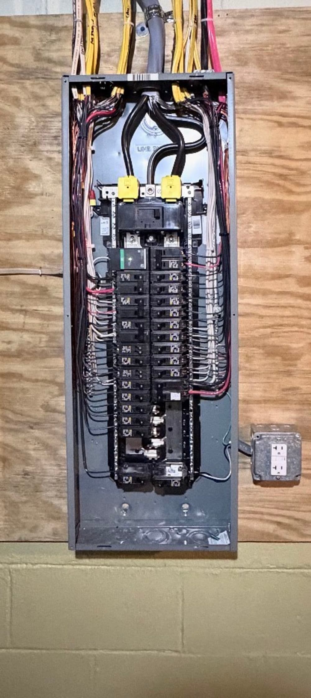 Electrical panel with circuit breakers and wiring, mounted on a wooden wall.