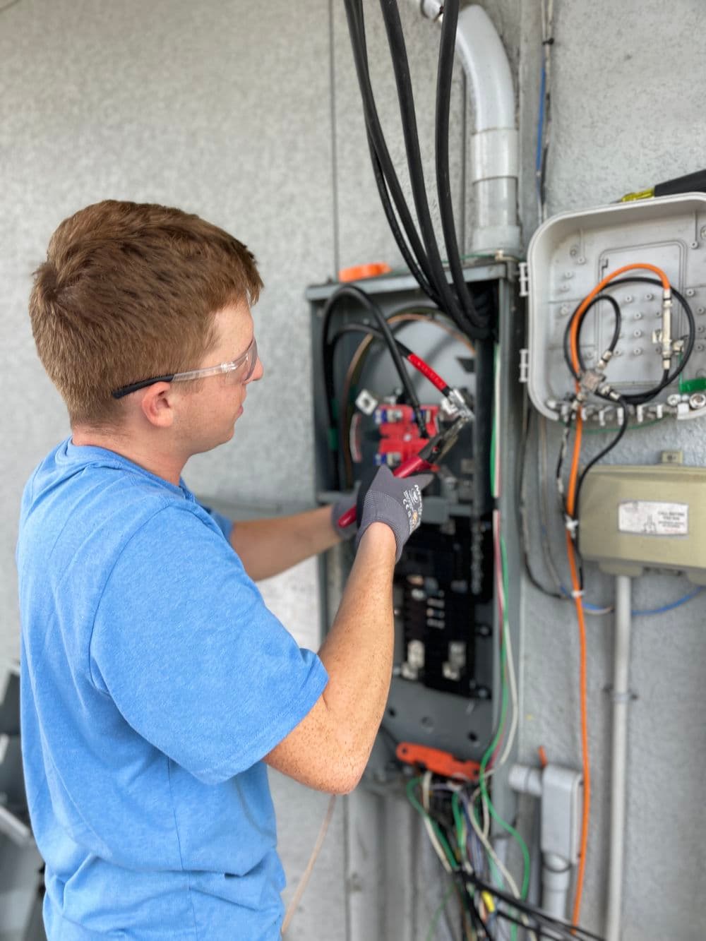 Electrician working on a circuit panel, wearing safety glasses and gloves, adjusting wiring.