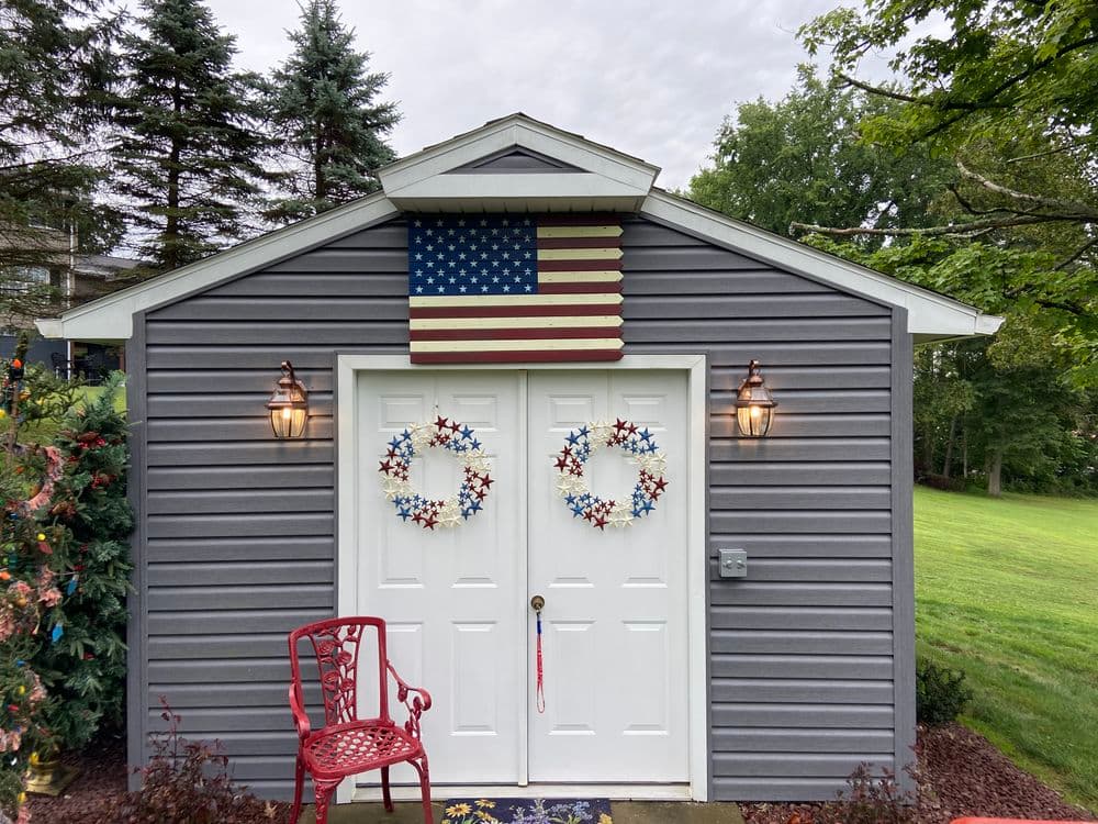 Gray shed with American flag, dual white doors, star-themed wreaths, and red chair.