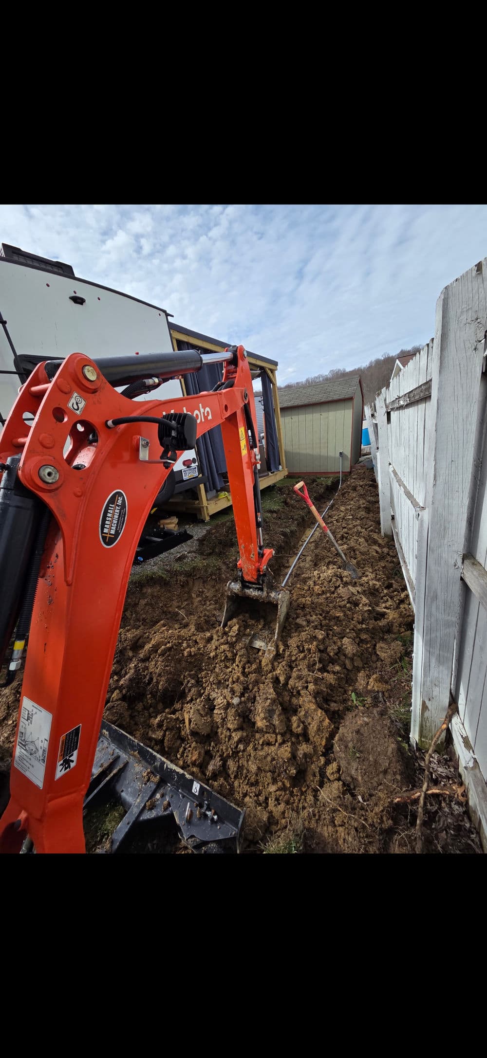 Excavator digging soil near wooden fence in residential area with cloudy sky.