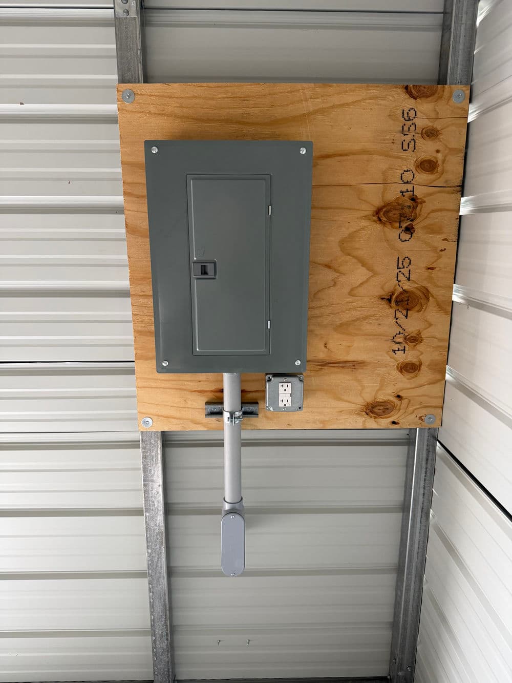Electrical panel mounted on wooden board inside a metal building, with a conduit and outlet.