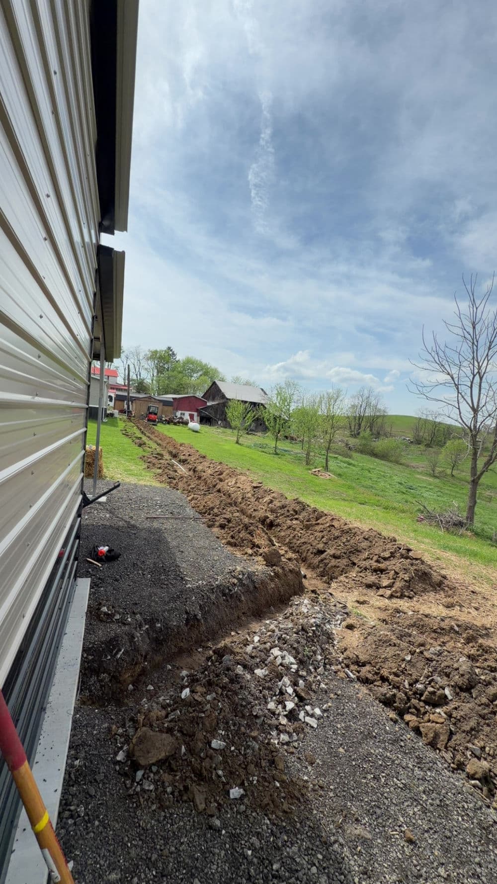 Excavation site beside a building with machinery, greenery, and distant barns in the background.