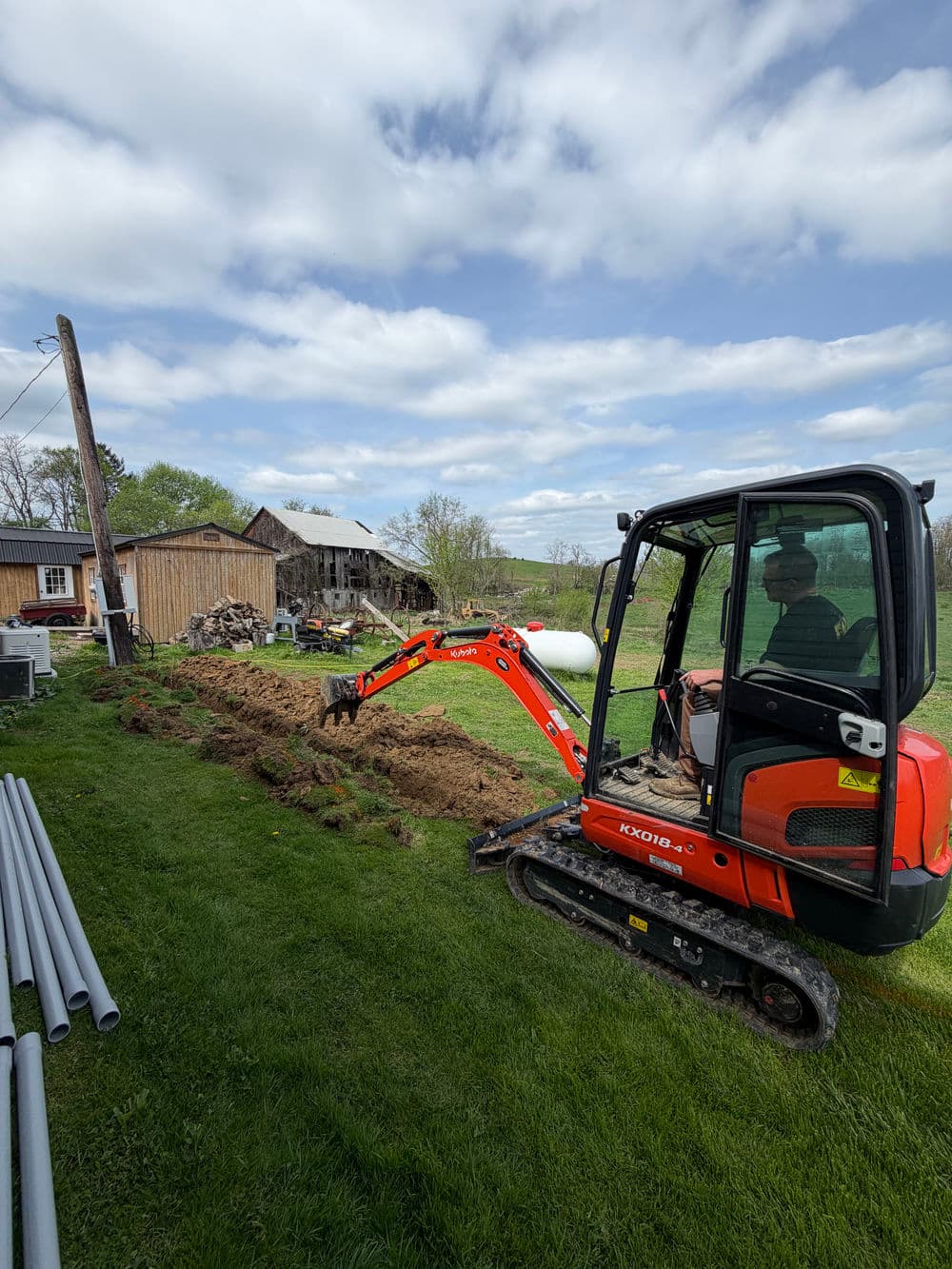 Excavator digging in yard near barn on cloudy day, with PVC pipes on grass.