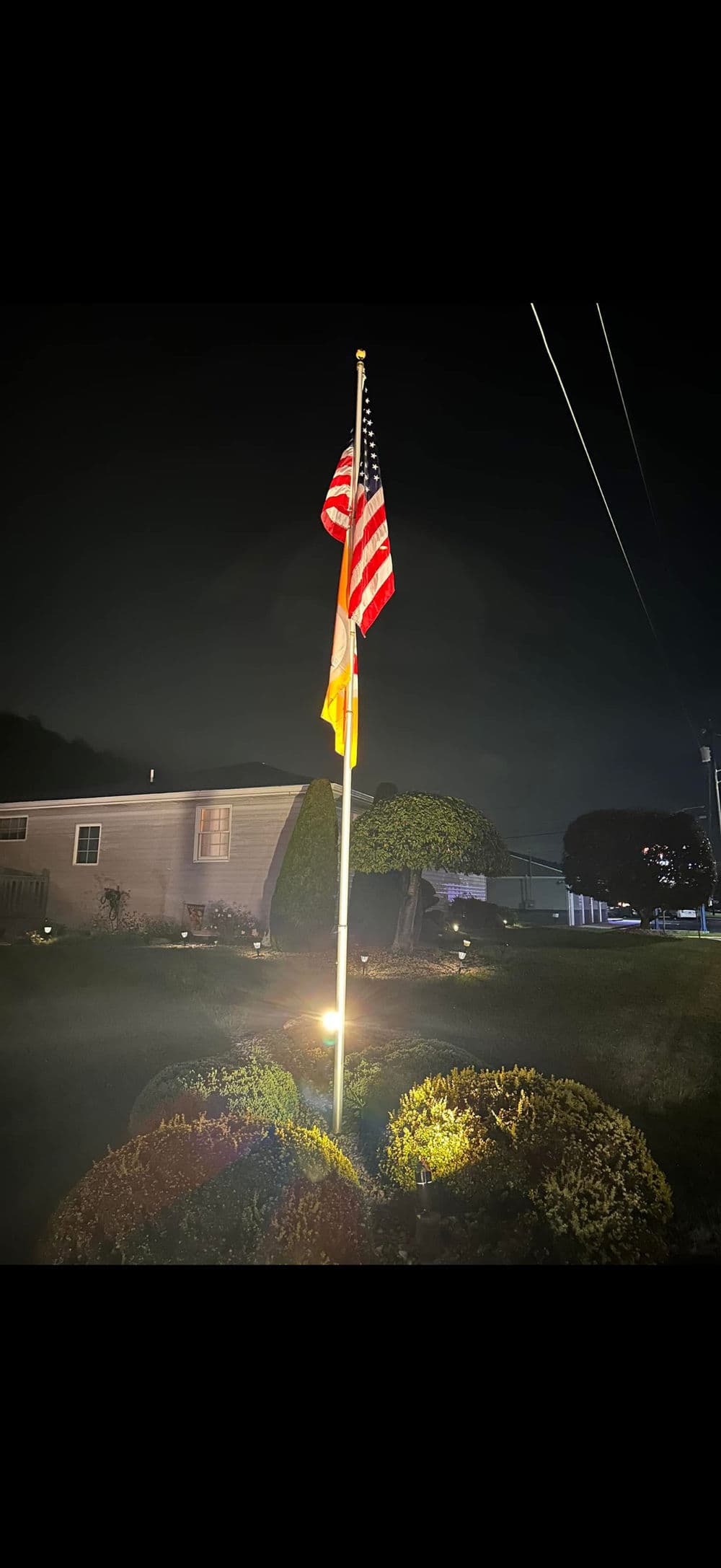 U.S. flag and a state flag lit at night in front of a house with landscaping.